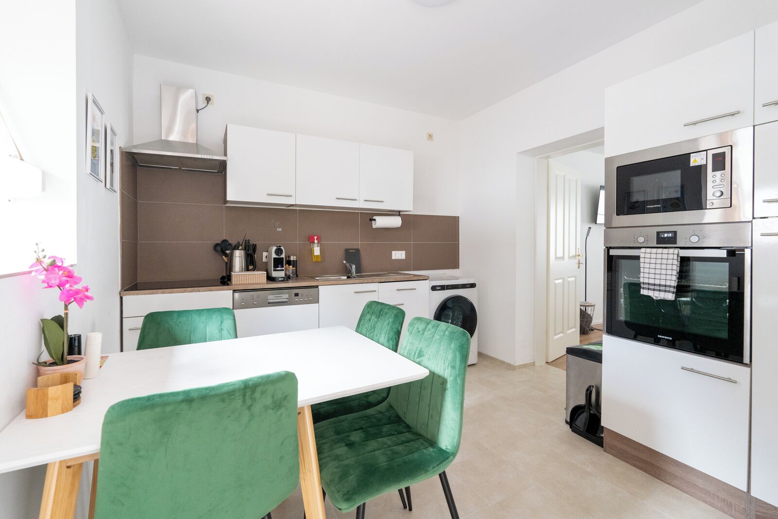 A modern kitchen with white cabinets, a built-in oven, a microwave, and a dishwasher. There is a dining table with green chairs.