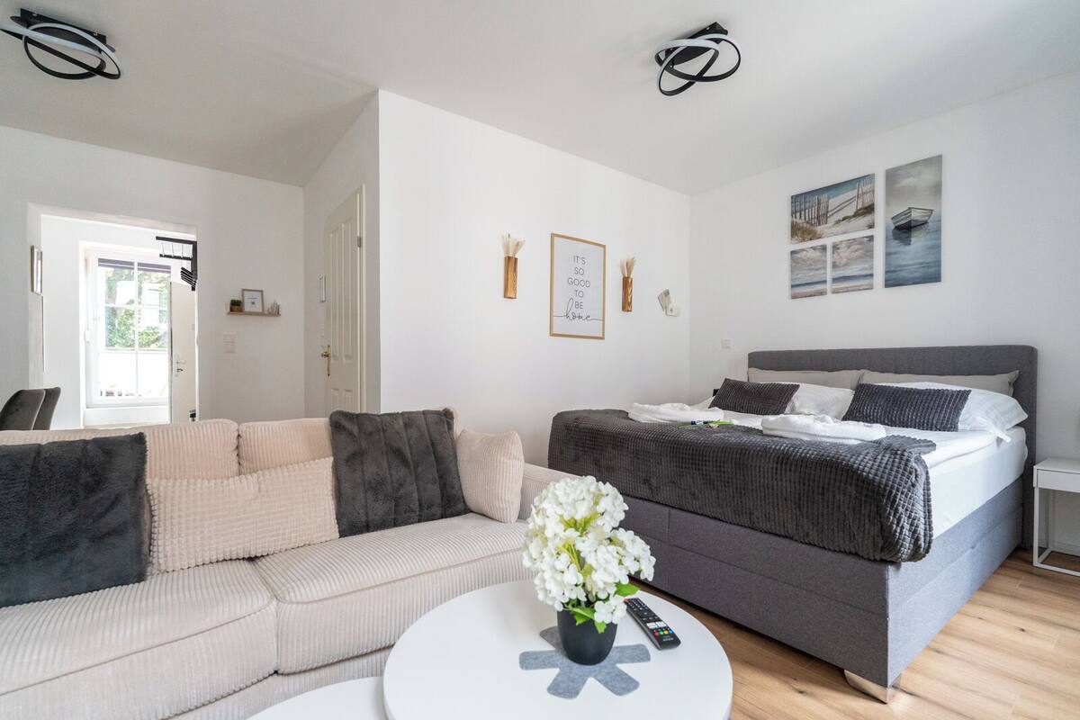 Ein modernes Schlafzimmer mit einem grauen Bett, einem beigen Sofa und einem weißen Couchtisch mit einer Blumenvase.