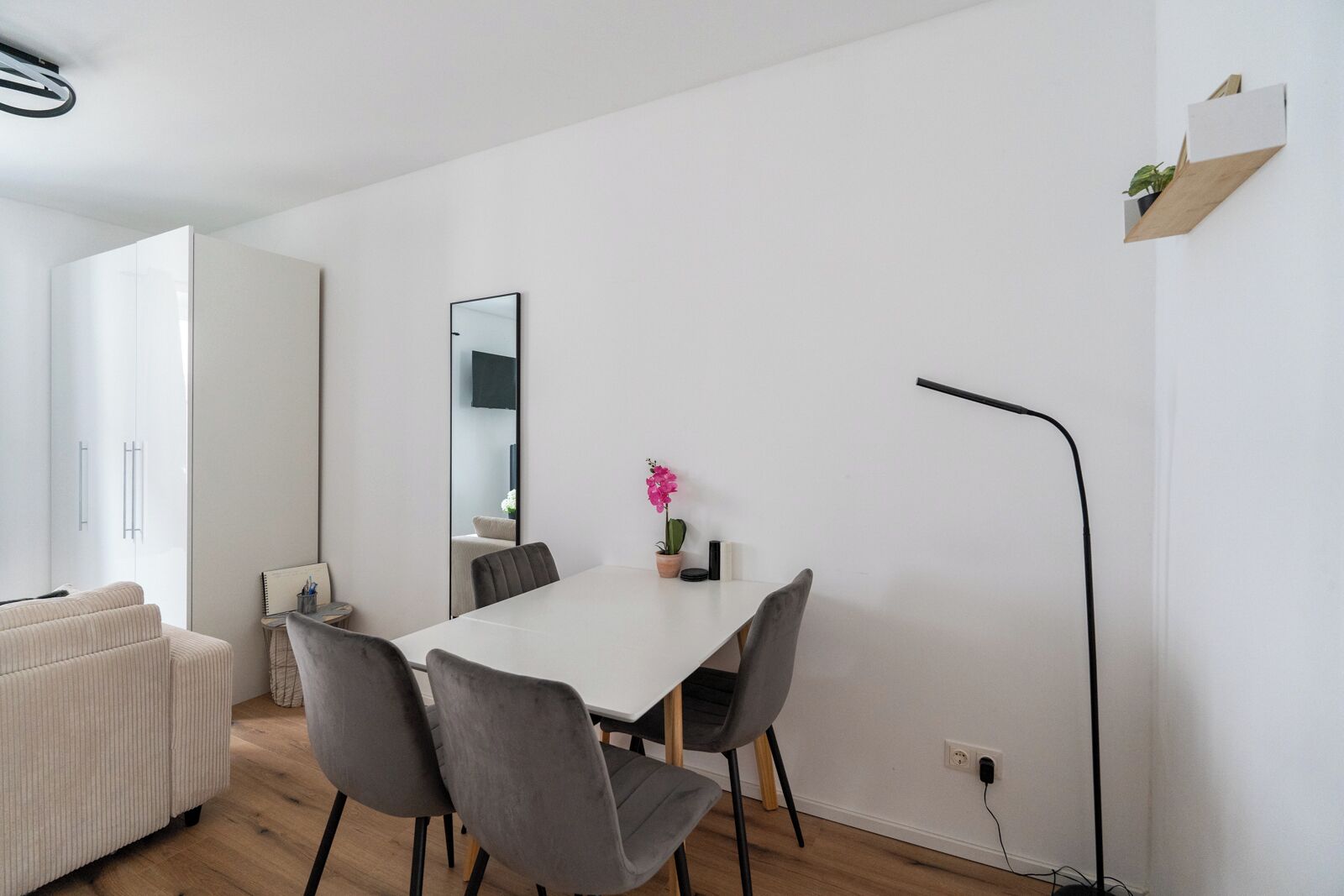 Una habitación moderna y minimalista con una mesa blanca, cuatro sillas grises, una pequeña planta en maceta y una lámpara de pie negra.