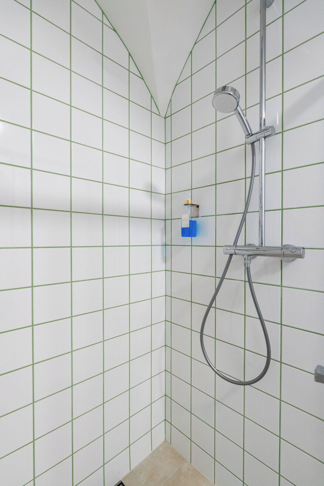 A shower with a handheld showerhead and a wall-mounted faucet, set against a tiled wall with a grid pattern.
