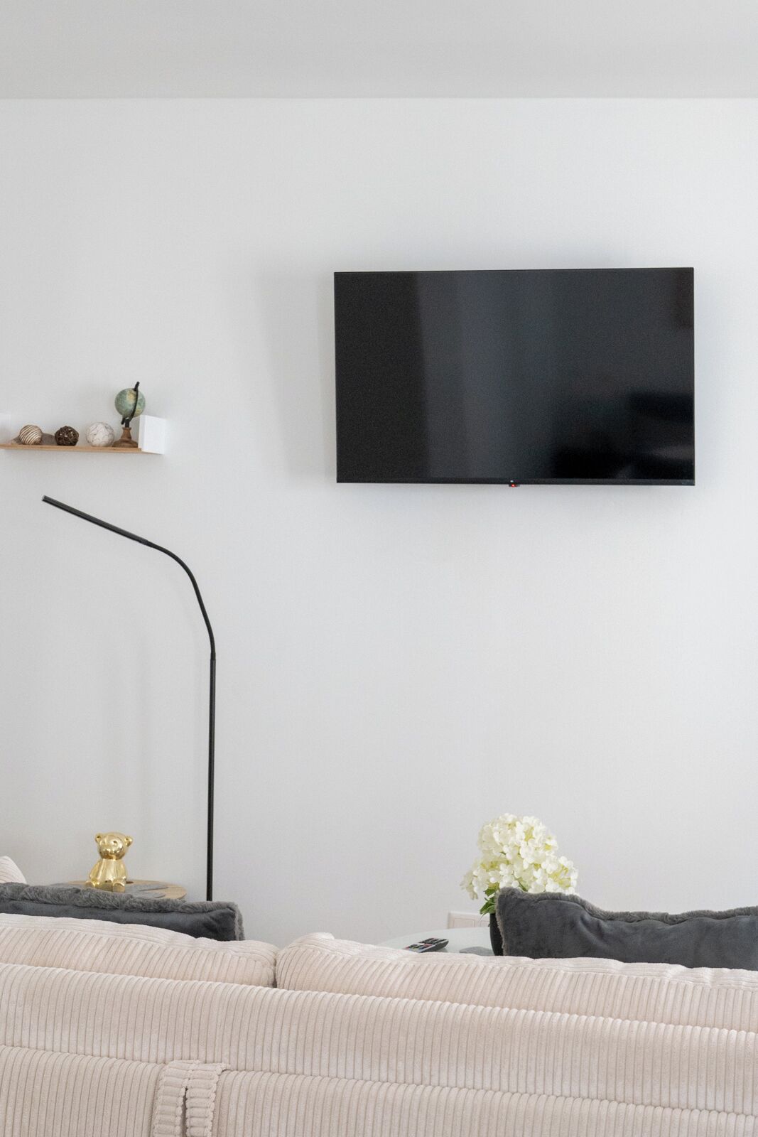 Un soggiorno moderno con una TV a schermo piatto montata al muro, un divano, un tavolino con una lampada e una piccola mensola con oggetti decorativi.