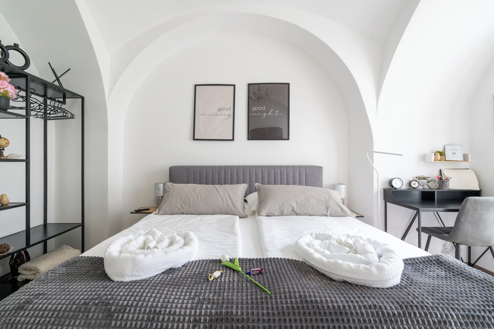 A modern bedroom with a grey upholstered headboard, two framed artworks, a desk with a chair, and a small shelf with decorative items.