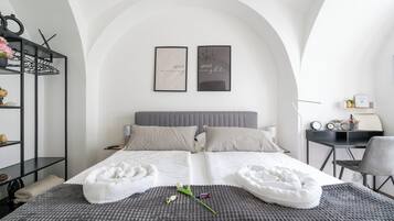 A modern bedroom with a grey upholstered headboard, two framed artworks, a desk with a chair, and a small shelf with decorative items.
