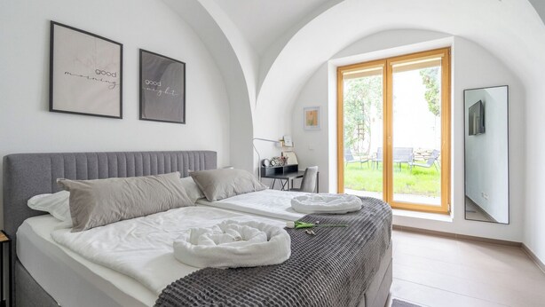 A bedroom with a bed, pillows, a bedside table, a desk, a chair, a TV, and a large window with a view of the outdoors. - COY Apartments Wachau - Relaxation In Style (Mautern an der Donau)