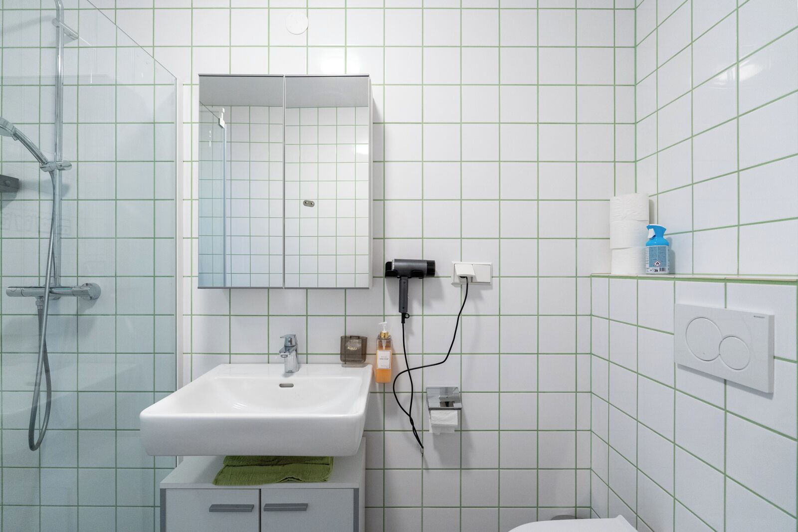 Ein Badezimmer mit einem weißen Waschbecken, Spiegel und Dusche. Es befinden sich Pflegeprodukte an der Wand und ein Handtuch auf der Ablagefläche.