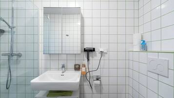 Ein Badezimmer mit einem weißen Waschbecken, Spiegel und Dusche. Es befinden sich Pflegeprodukte an der Wand und ein Handtuch auf der Ablagefläche.
