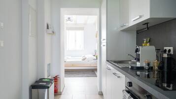 Un corridoio stretto conduce a una camera da letto con un letto e una finestra. Sulla destra si vede un angolo cottura con lavello, fornelli e diversi oggetti da cucina.