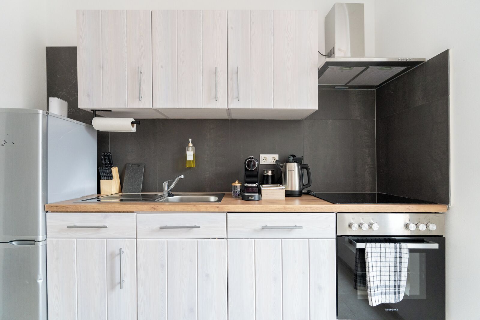 A modern kitchen with light wood cabinets, a stainless steel refrigerator, a coffee maker, and a kettle on the counter.