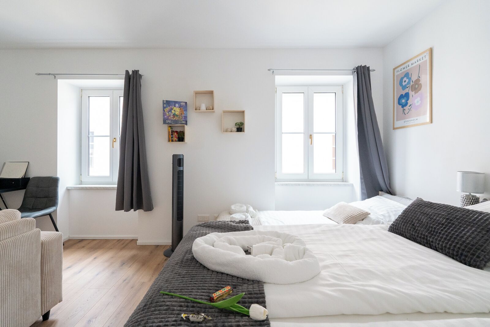 A modern bedroom with a large bed, two windows with curtains, a standing heater, a desk with a chair, and a small shelf with decorative items.
