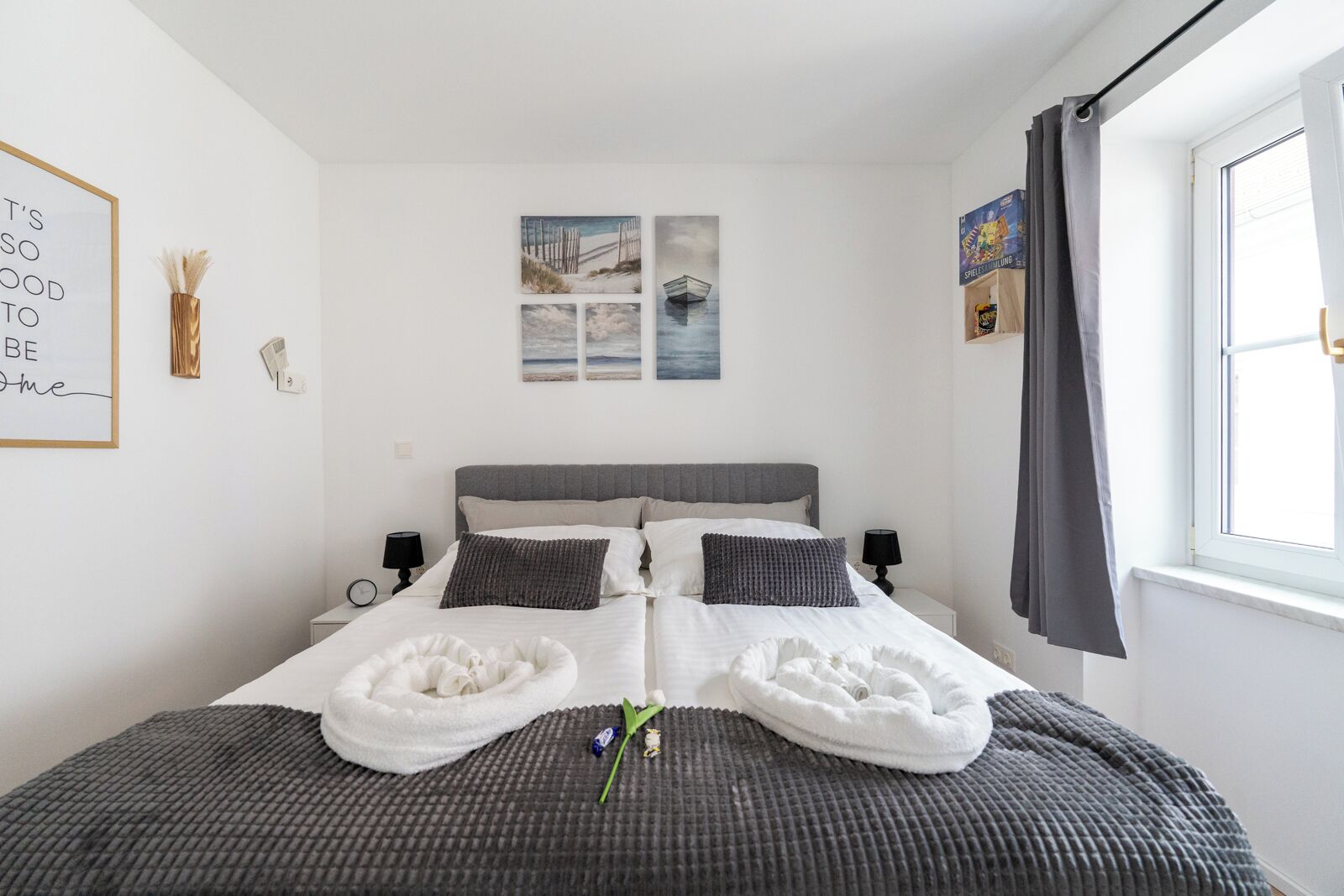 En pænt redt seng med hvidt og gråt sengetøj, to sengeborde med lamper, et vindue med gardiner og vægkunst bestående af et indrammet citat og et strandmotiv.