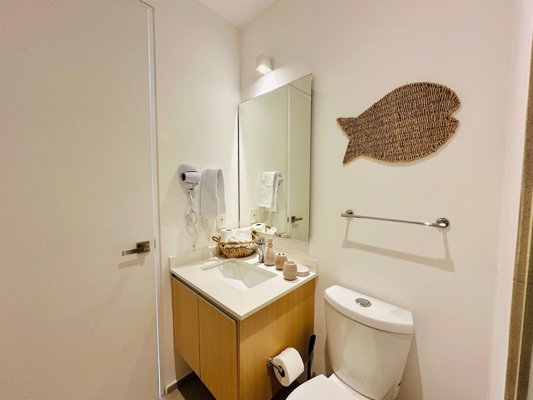 A bathroom with a toilet, sink, and a mirror. There is a towel rack, a hairdryer, and a basket with toiletries.