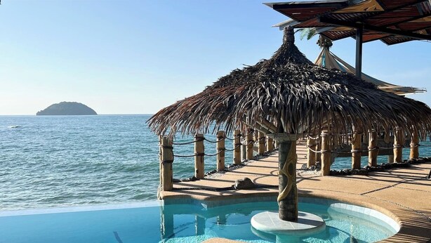 Zona de piscina con sombrilla de paja, bañera de hidromasaje y vista al mar.