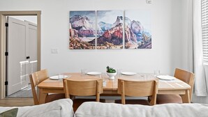 A dining area with a wooden table and chairs, a small plant centerpiece, and a painting of mountains on the wall.