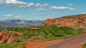 A golf course with a road curving along the edge, surrounded by red rock formations and distant mountains.