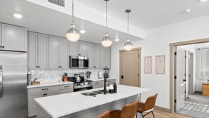 A modern kitchen with grey cabinets, stainless steel appliances, a central island with bar stools, pendant lighting, and a herringbone tile backsplash.