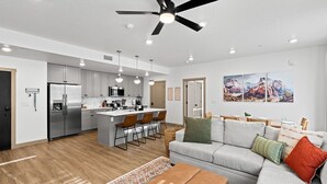 A modern kitchen with stainless steel appliances, a central island with bar stools, a ceiling fan, recessed lighting, and a dining area with a table and chairs.