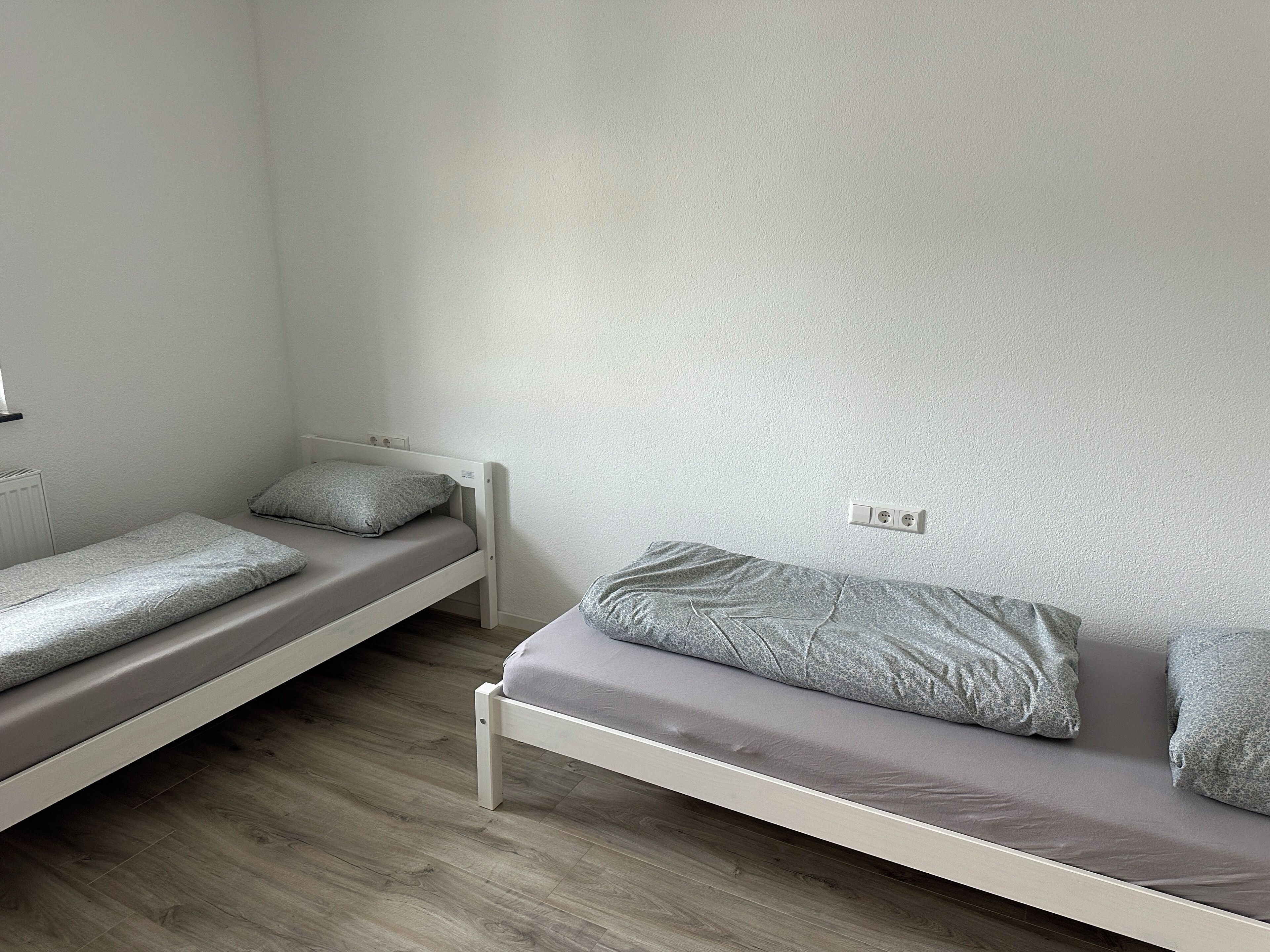 Ein Zimmer mit zwei Einzelbetten, jeweils mit grauem Lattenrost und passenden Kissen, sowie einem weißen Kopfteil.