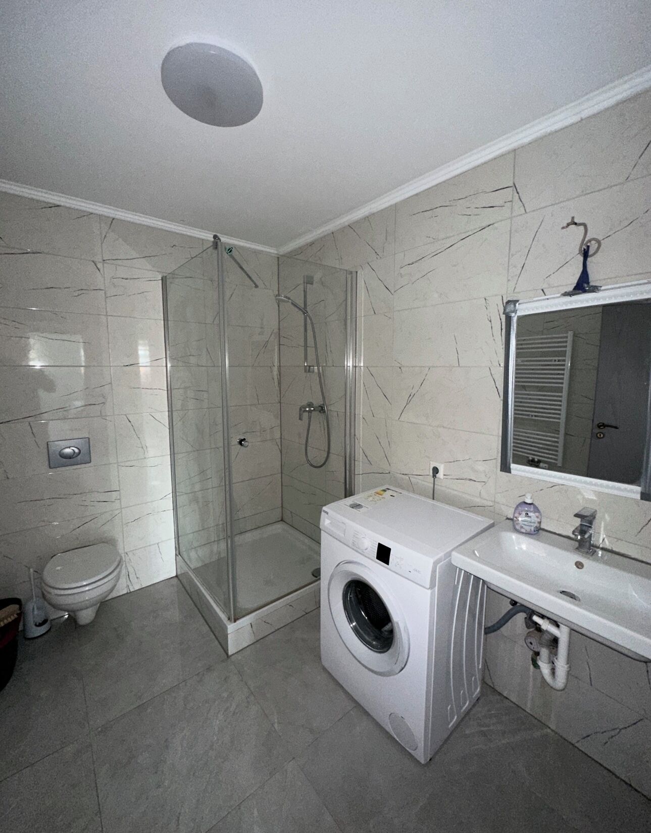 Casa de banho com cabine de chuveiro em vidro, máquina de lavar roupa, sanita e lavatório com espelho.