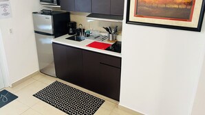 A compact kitchen with dark cabinets, a microwave, a coffee maker, and a sink. There is a framed picture on the wall and a doormat on the floor.