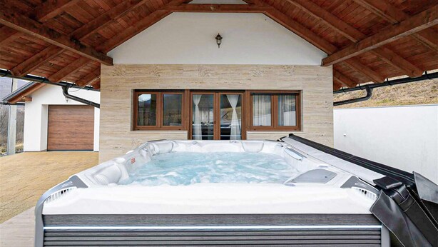 A hot tub under a wooden roof with a stone wall and windows.
