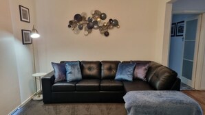 Un salon avec un canapé en cuir noir, un pouf gris et un élément décoratif fixé au mur.