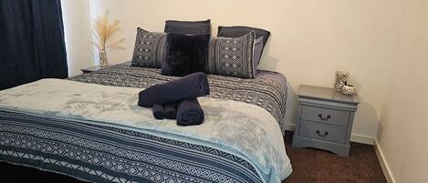 A neatly made bed with patterned bedding, a bedside table with a lamp, and a small vase with dried plants.