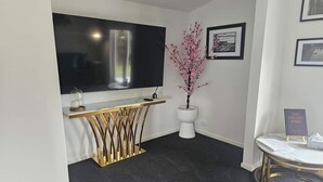 Un salon moderne avec un grand téléviseur à écran plat fixé au mur, une console dorée, un vase blanc contenant des fleurs roses et une petite table ronde sur laquelle est posé un livre.