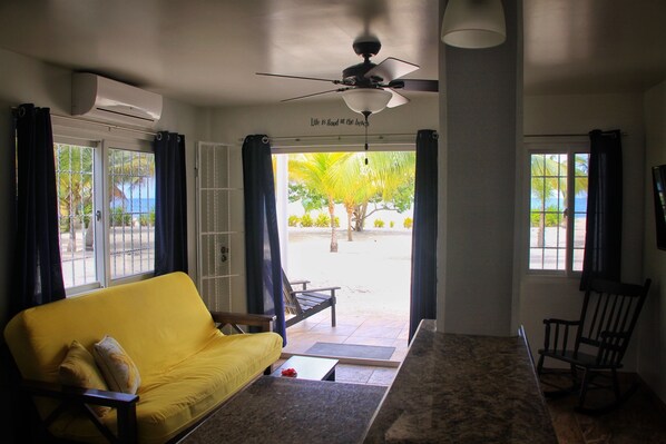 A room with a yellow sofa, a ceiling fan, and a view of a palm tree and beach through the door.