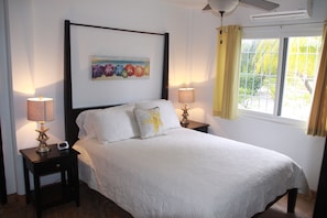 A neatly made bed with white linens, two bedside tables with lamps, a painting above the bed, and a window with curtains.