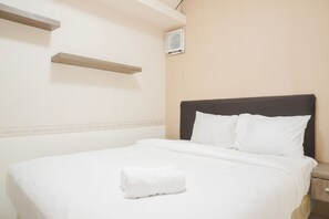 A neatly made bed with white linens and a rolled white towel, a bedside table, and a wall-mounted air conditioner. - Comfort And Tidy 2Br At Green Bay Pluit Apartment Near Mall (Jakarta)