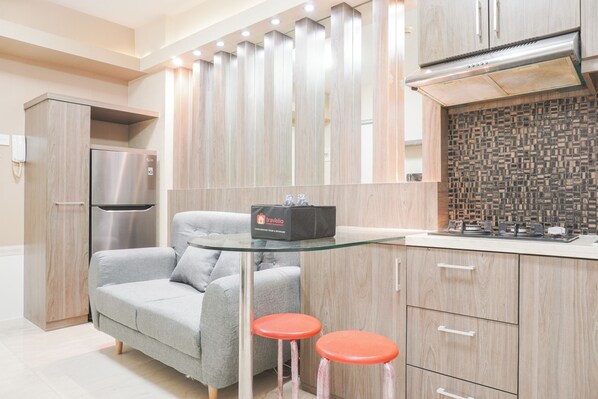A modern kitchen with a grey sofa, two red stools, a stainless steel refrigerator, and a travel bag on a glass table. - Comfort And Tidy 2Br At Green Bay Pluit Apartment Near Mall (Jakarta)