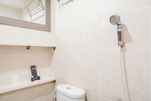 A bathroom with a toilet, a shelf with toiletries, and a shower head. - Comfort And Tidy 2Br At Green Bay Pluit Apartment Near Mall (Jakarta)