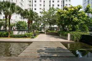 A pathway leading to a building with multiple floors, surrounded by lush greenery and palm trees. - Comfort And Tidy 2Br At Green Bay Pluit Apartment Near Mall (Jakarta)