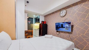 Ett hotellrum med en säng, en tv som är monterad på väggen, en klocka och ett litet kök i bakgrunden.
