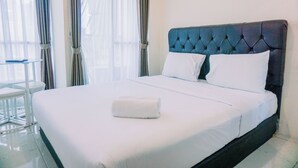 Un lit bien fait, avec du linge de lit blanc et une serviette blanche pliée, une tête de lit rembourrée de couleur foncée et une petite table ronde placée à côté du lit.