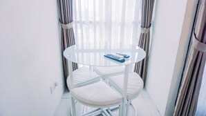 Une petite table ronde et blanche, sur laquelle se trouvent un coussin et une télécommande, placée dans un coin, avec des rideaux et une fenêtre en arrière-plan.