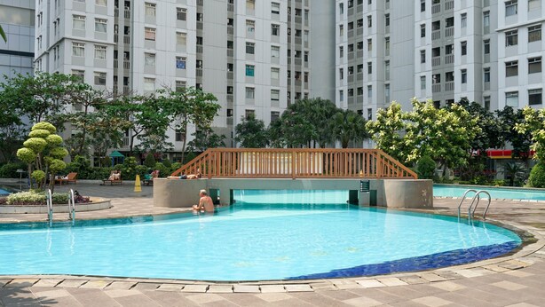 A swimming pool with a wooden bridge, surrounded by greenery and a tall residential building in the background.