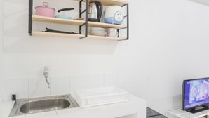 A kitchen with a stainless steel sink, a white countertop, a wooden shelf with kitchenware, and a television on a stand.