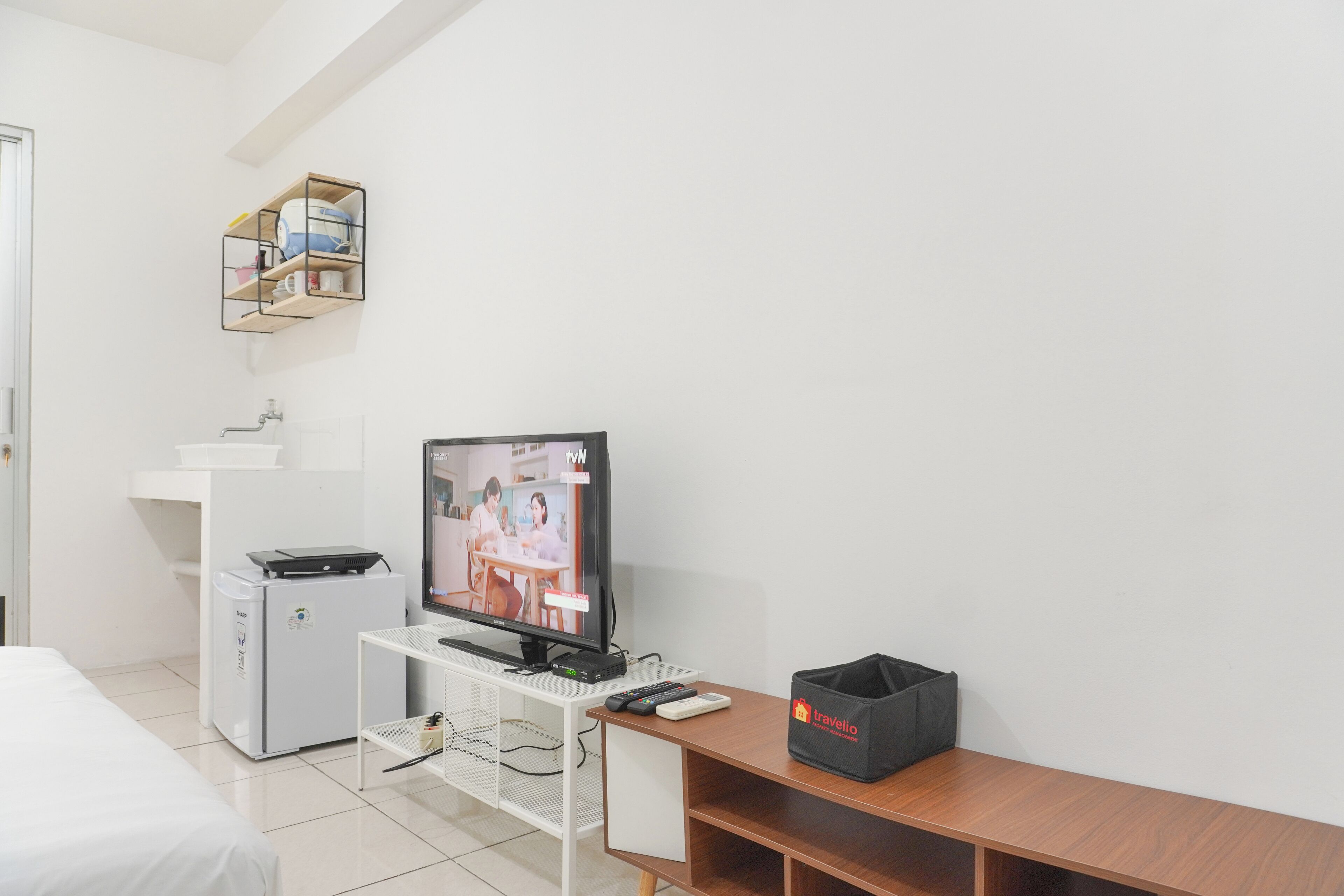 A hotel room with a bed, a desk with a computer, a TV, a small refrigerator, a shelf with toiletries, and a trash bin.