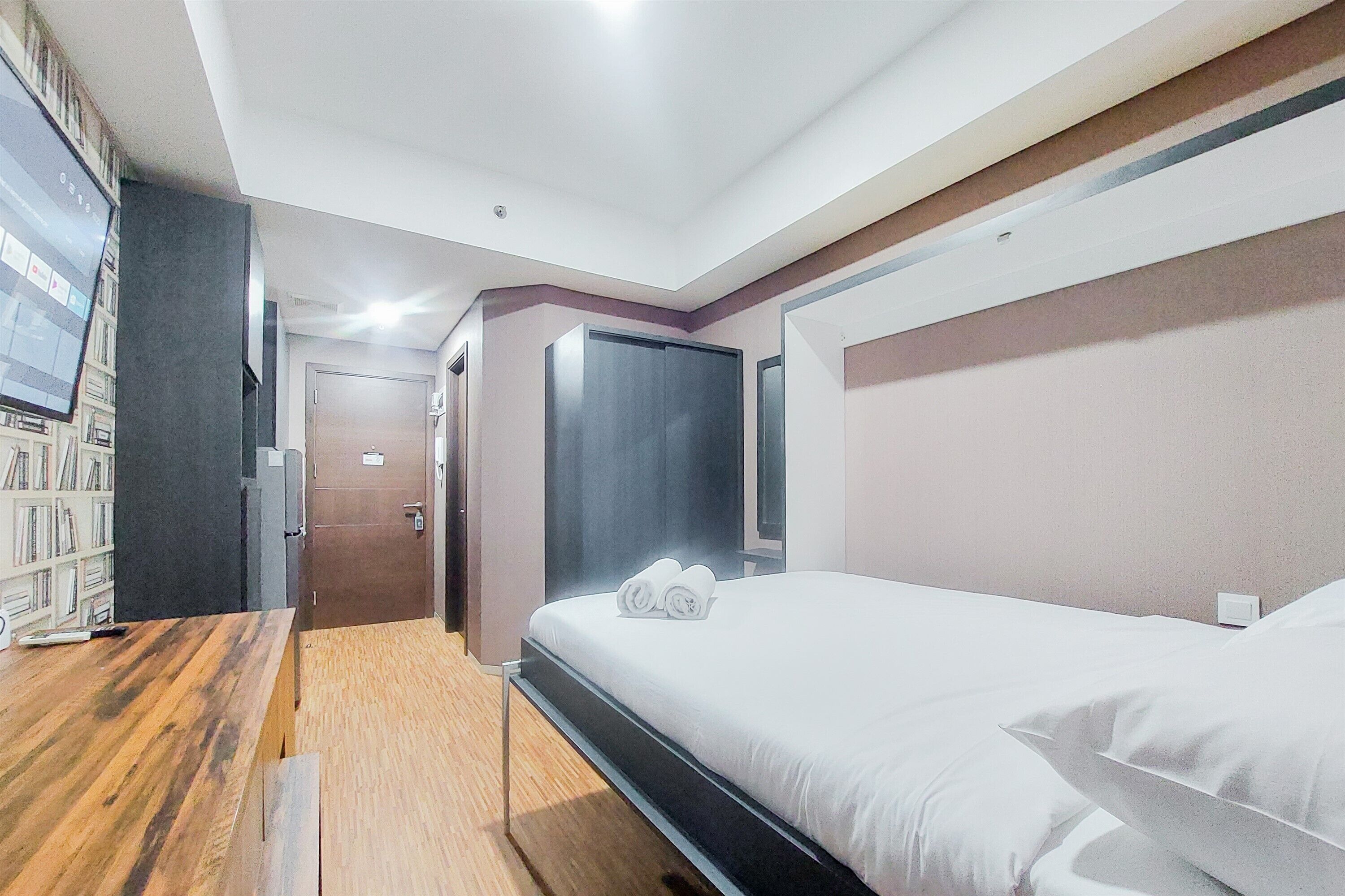 A modern hotel room with a wooden desk, a bed with white linens, a flat-screen TV, and a bookshelf.