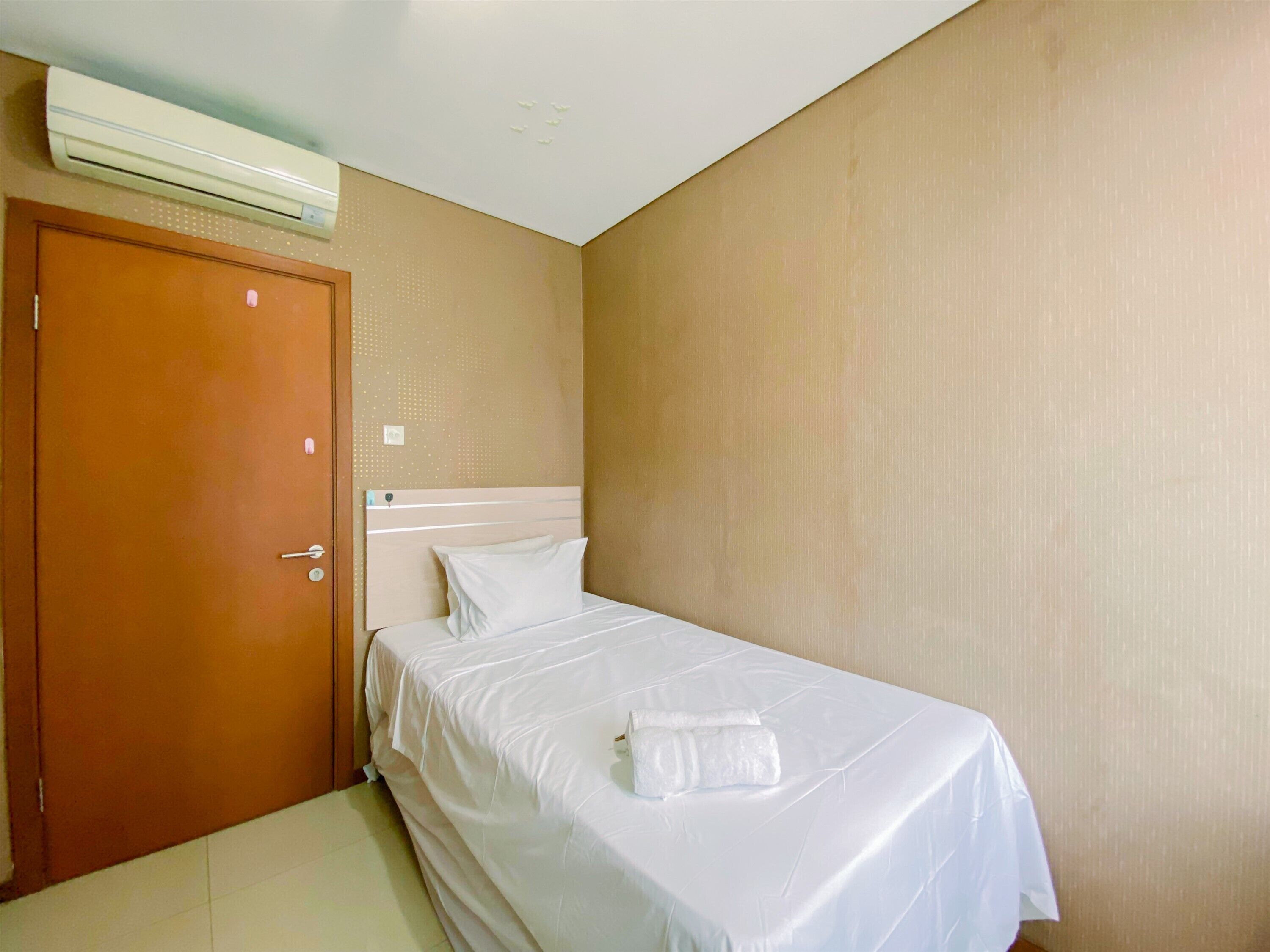 Un lit simple avec du linge de lit blanc et une serviette, une porte en bois et un appareil de climatisation fixé au plafond.
