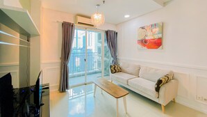 A modern living room with a sofa, a coffee table, and a TV. There is a balcony with curtains and a view of buildings. - Comfort Living 3Br Apartment At 25Th Floor Thamrin Residence (Jakarta)