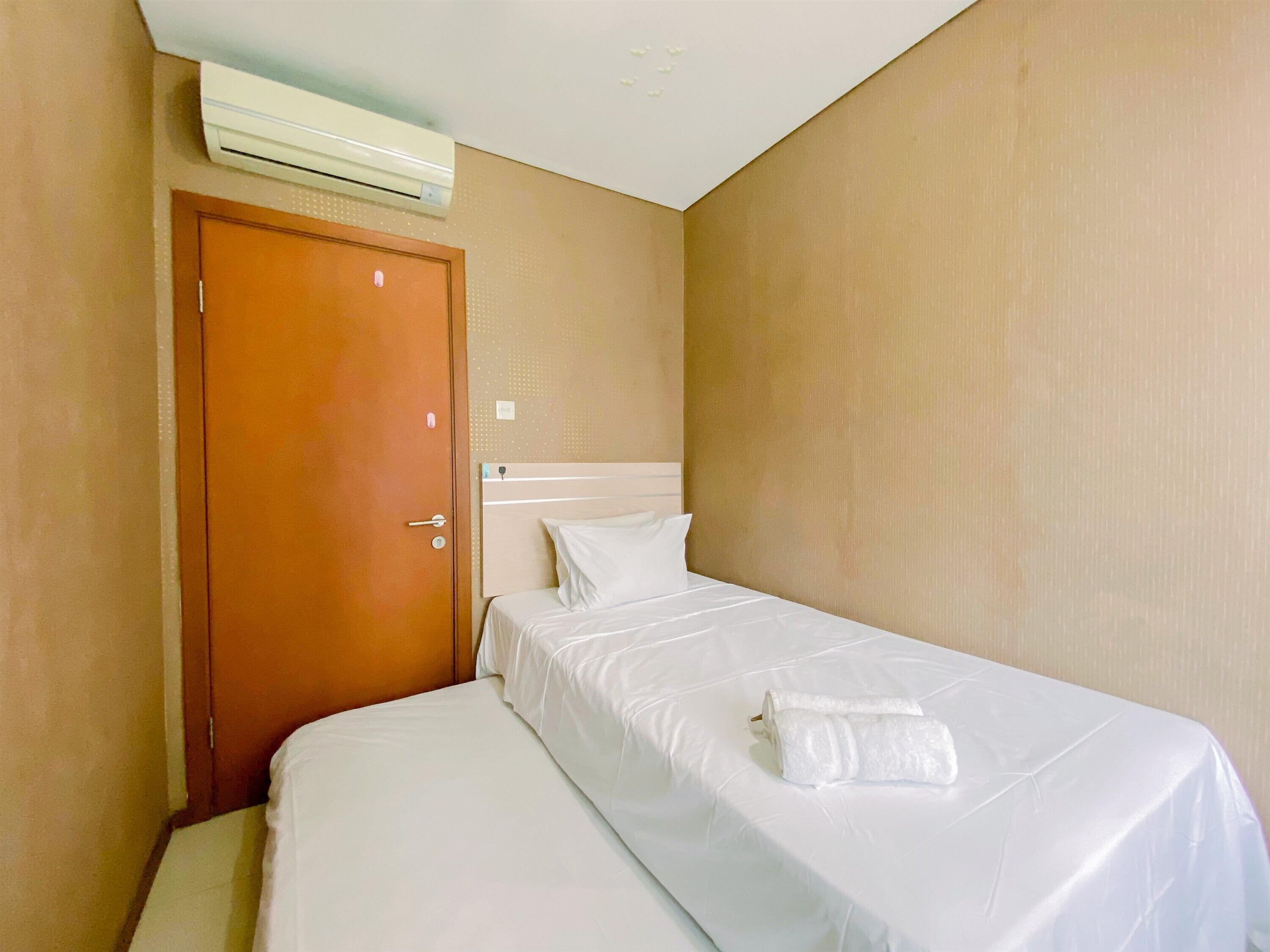 A single bed with white linens and a folded towel, a wooden door, and an air conditioning unit on the wall.
