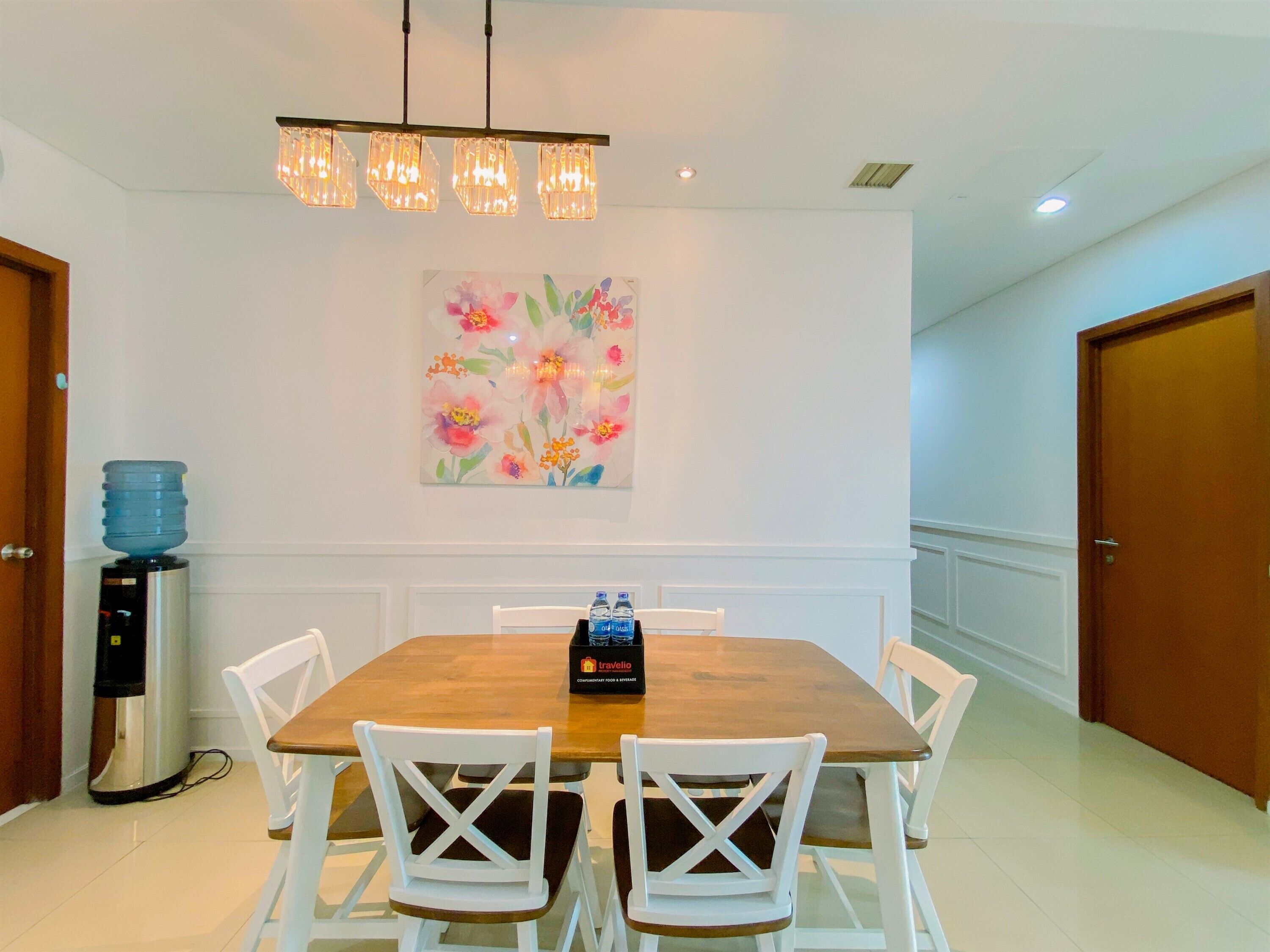 A dining area with a wooden table, white chairs, a chandelier, a water dispenser, and a travel-themed centerpiece.