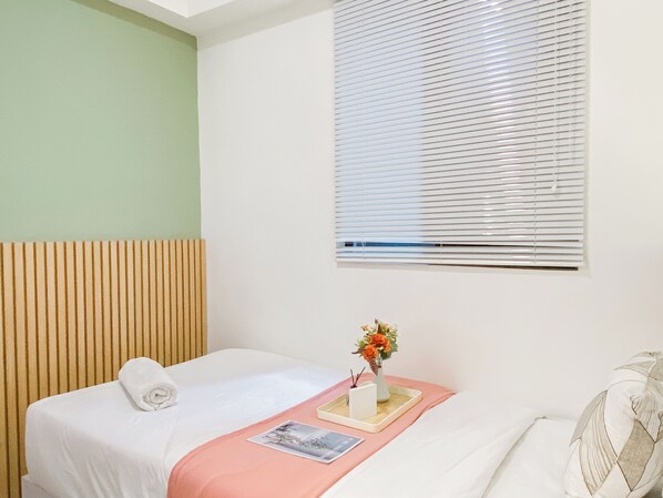 A hotel room with a bed, a window with blinds, a bedside table with a vase of flowers, and a wall with a vertical wooden panel. - Restful And Homey 2Br At Meikarta Apartment (Cikarang)