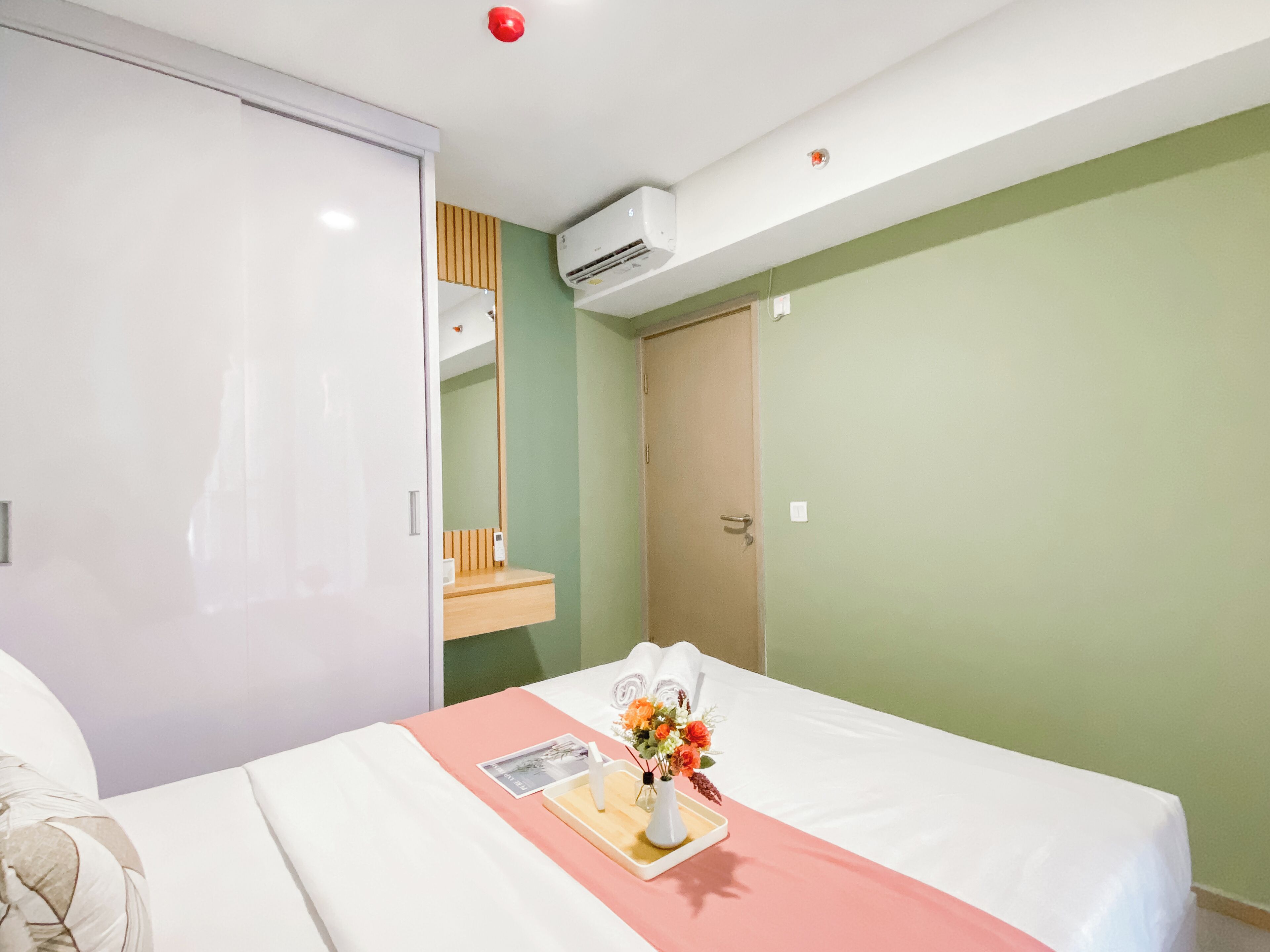 A hotel room with a bed, a bedside table with a vase of flowers, a mirror, an air conditioning unit, and a door.