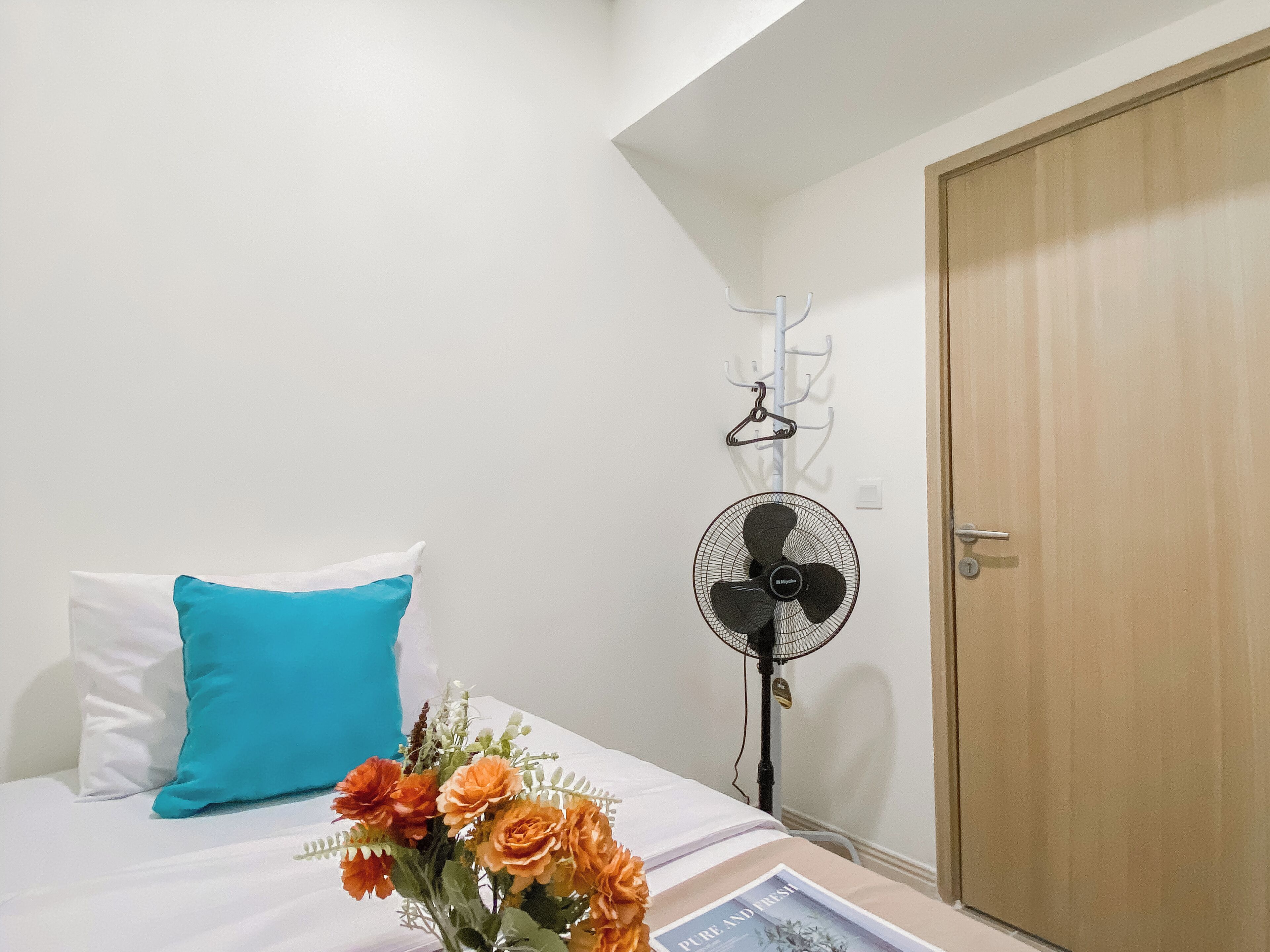 A bedroom with a bed, a blue pillow, a white wall, a wooden door, a standing fan, and a wall-mounted coat rack.