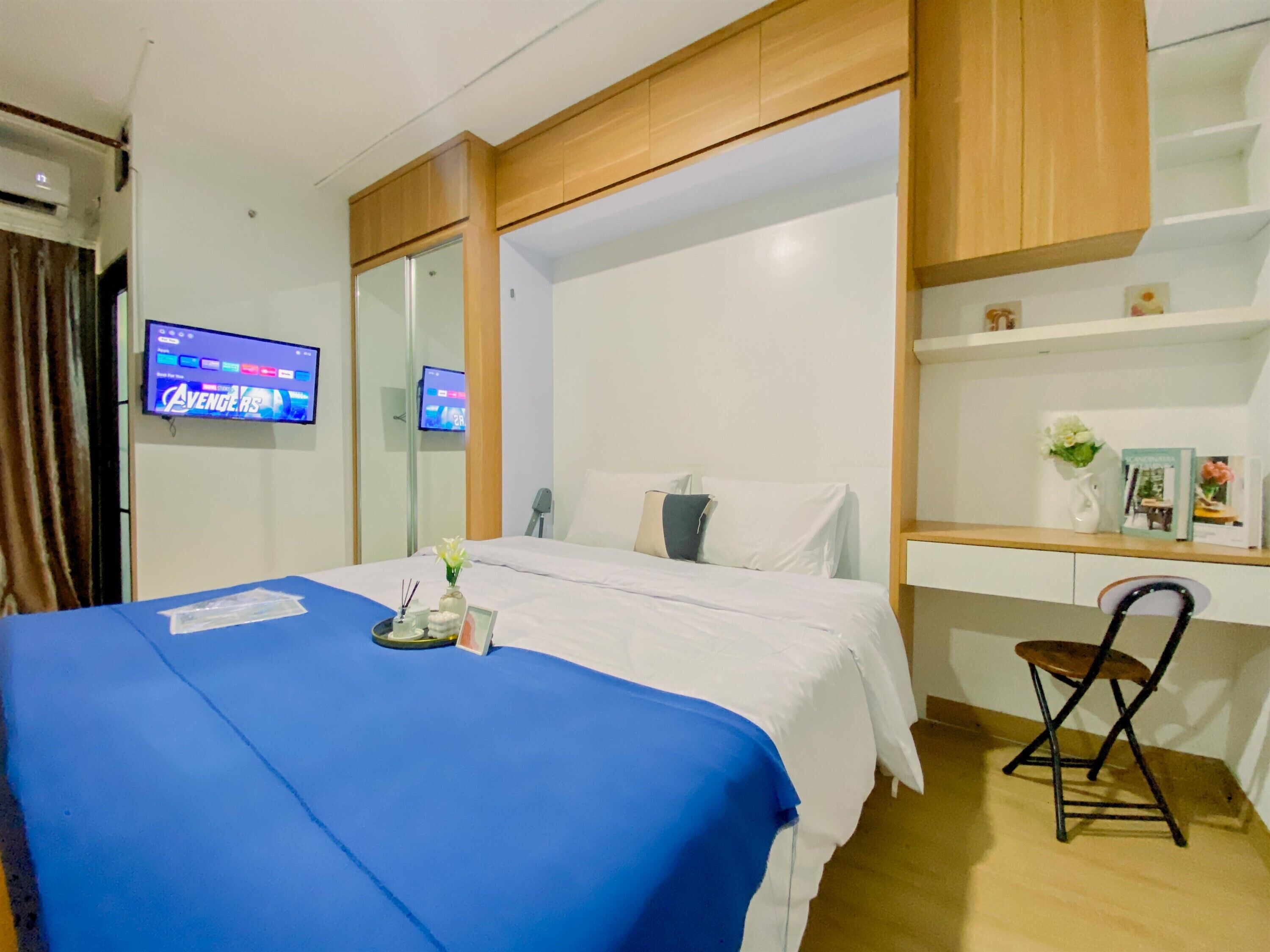 Quarto de hotel com cama, escrivaninha, cadeira, televisão e prateleira com objetos decorativos.