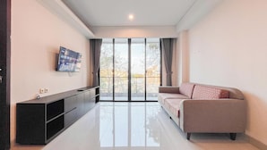 A modern living room with a flat-screen TV, a sofa, and a glass door leading to an outdoor area. - 2BR Apartment at Skales Residence Near Nusa Dua Square (Nusa Dua)
