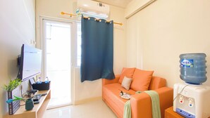 A small, well-lit room with a sofa, a water dispenser, a TV, and a desk with a computer and a plant. - Good Choice And Restful 2Br Green Pramuka City Apartment Near Mall (Jakarta)